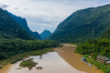 A broad muddy river curves between steep, forested mountains under a bright sky in northern Laos. Riverside vegetation, longboats, and dense green hills create a dramatic tropical landscape.