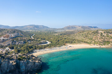 Aerial perspective of Vai beach in Crete, featuring a unique palm forest bordering a golden sandy bay and clear turquoise water. Rugged hills and a winding coastal road complete the sunlit