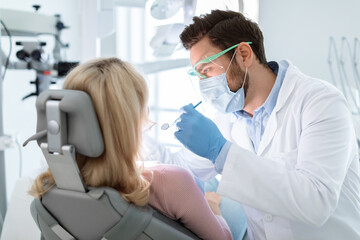 Man dentist in face mask and glasses doing treatment for patient blonde lady, holding dental tools,...