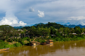 Obraz premium Traditional wooden boats moored along a lush green riverbank on the Mekong River in Luang Prabang, Laos. The scene features tropical trees, distant hills, and dramatic cumulus clouds under bright