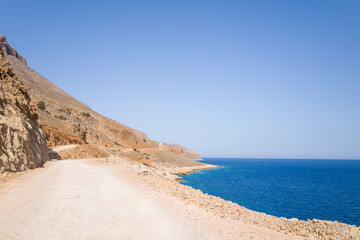 Sun-bleached unpaved road hugs rugged ochre cliffs above the deep blue Mediterranean Sea near Balos, Crete. The landscape feels wild, open, and bathed in clear daylight.