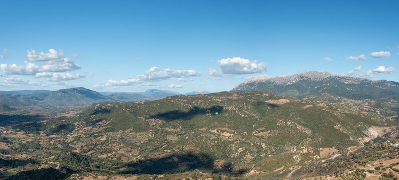 Sweeping view of rugged hills and distant mountain peaks near Orgosolo, Sardinia, under a clear blue sky dotted with fluffy clouds. Sunlight highlights the varied terrain and natural textures of the - Powered by Adobe