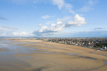 Aerial view of Ouistreham reveals a sprawling sandy shoreline at low tide, with the coastal town stretching along the horizon beneath a bright blue sky dotted with clouds.