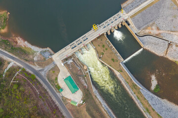 Top-down image displays a modern hydroelectric dam spanning a calm reservoir, with water released through spillways into a controlled channel. The engineered embankments, access road, and sparse
