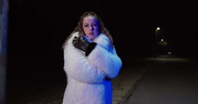 Cinematic night shot of a woman alone on a quiet street, fastening her coat and cuddling into it for warmth, looking sad and abandoned. Concept for loneliness, breakup, winter cold, emotional
