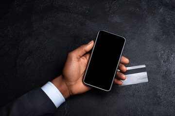 Hand holding smartphone with blank screen and credit card on dark background. Mobile payment, online shopping, fintech and digital banking concept.