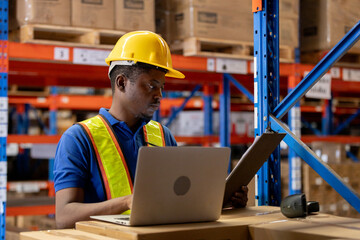 Focused warehouse worker using laptop and clipboard for inventory management, data entry in storage area, representing modern logistics, efficiency, digital supply chain operation.