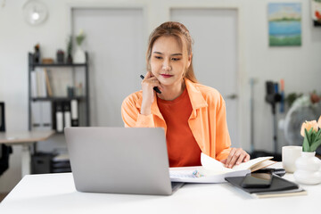 Concentrated Asian businesswoman working on laptop and taking notes at home office desk, focused professional managing project tasks online.
