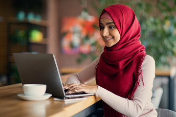 Cheerful young middle-eastern woman in hijab using laptop while drinking coffee at cafe, female...