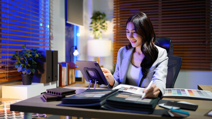 Asian Interior Designer at Work Selecting Color Palette and Material Samples for Home Design Project Online Using Laptop in Modern Office Space at night