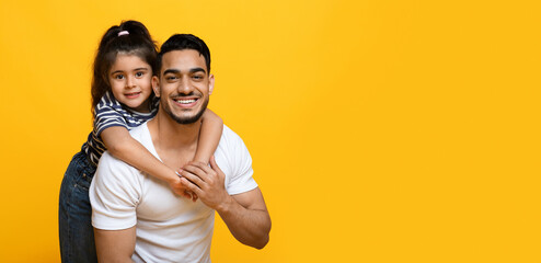 Fototapeta premium Portrait Of Happy Young Middle Eastern Dad And Cute Little Daughter Posing Together Over Yellow Background, Female Kid Embracing Daddy From The Back And Smiling At Camera, Panorama, Copy Space