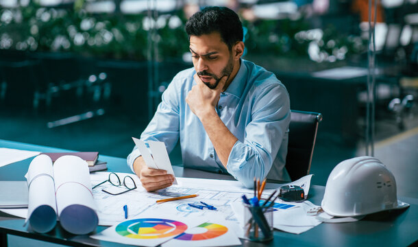 A man sits at a desk in a modern office, looking at project plans with a thoughtful expression. Various documents, blueprints, and a hard hat spread around him. It is daytime.