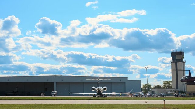 Helicopter flying near the runway and air traffic control tower at a busy airport while several airplanes are parked outside hangar. 