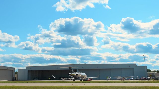 A private jet is parked on the airport apron as a ground crew worker prepares to tow aircraft near a large hangar on a sunny day.