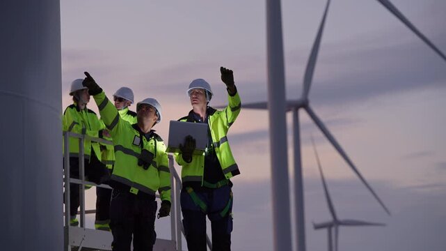 Engineers wearing safety helmets and jackets inspecting a wind turbine at wind farm at sunset. Concept renewable energy, sustainable technology wind power maintenance and environmental engineering.
