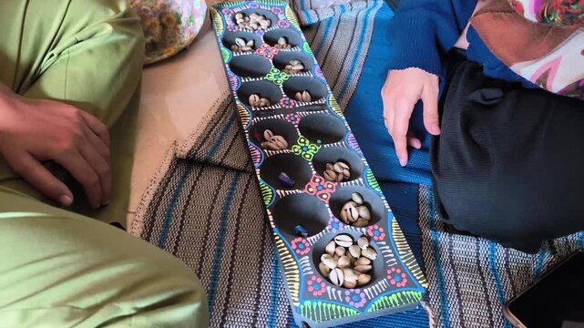 Two people playing a traditional board game known as Congklak or Dakon, using seashells.