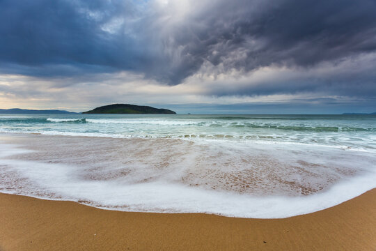 Dramatic storm clouds and dramatic lighting over waves washing onto a sandy beach