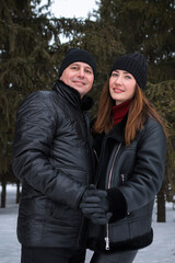 Pair Dressed Warmly Beside Evergreen Background, Two Individuals Wearing Coats And Hats In Urban Setting, Couple Exuding Fashion In Winter Attire With City Vibe And Cozy Atmosphere