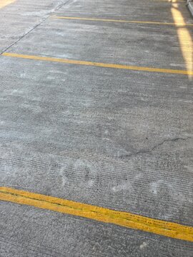 High-angle view of an empty concrete parking lot or paved industrial surface. It features parallel yellow painted lines and a textured, weathered gray ground.