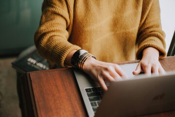 Close-up of person in yellow sweater typing on laptop beside journal, bracelet visible, symbolizing study, work, and creativity.