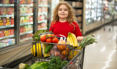 Child shopping with cart in supermarket. Kid choosing fresh vegetables in grocery store. Child with food in food store. Kid selecting produce at grocery store. Child with fresh food in grocery shop.