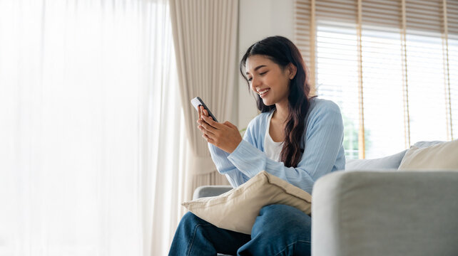 Smiling pretty young Asian woman sitting on cozy couch, using funny mobile apps in living room. Woman at Home, Doing Online Shopping, Messaging Friends, Posting on Social Media