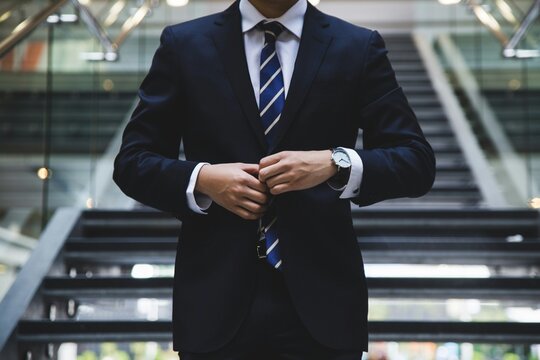 Close-up of man in dark suit adjusting jacket near staircase with glass railings, symbolizing professionalism and corporate lifestyle. - Powered by Adobe