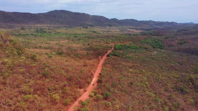 Drone tracks 4x4 penetrating red-soil road through cerrado woodland with rocky ridge escarpment forming dramatic background, red latosoil exposed, dry season in Tocantins central plateau.
