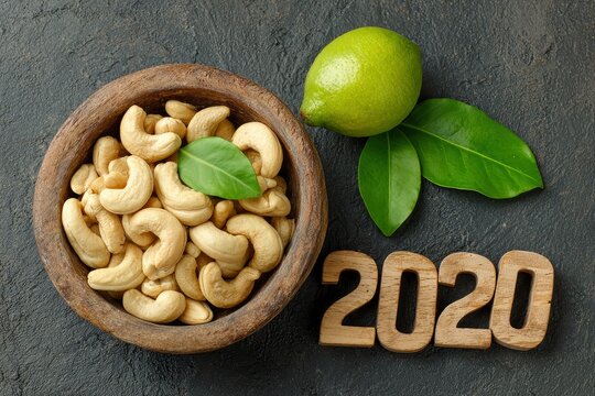 An appealing overhead shot showcases a rustic wooden bowl overflowing with numerous creamy, raw cashew nuts, complemented by a vibrant fresh green leaf. Adjacent to the bowl, a whole lime adds a splas