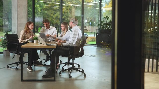 A view through a glass partition shows a modern, busy office interior. Business people are working at desk