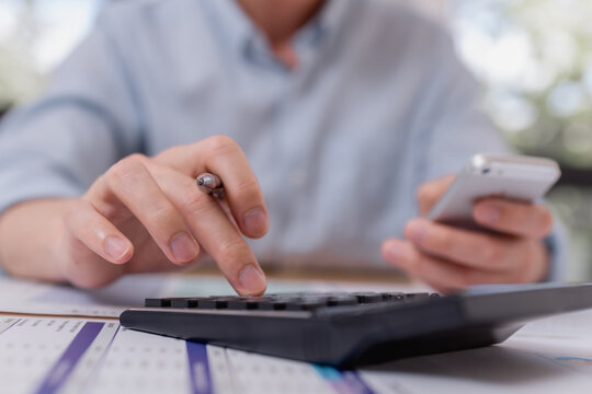 businessman using calculator and smartphone while analyzing financial data on paper documents, focusing on expense tracking and budgeting in office workspace. - Powered by Adobe