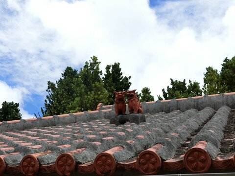 Two traditional Shisa statues on a red tile roof under a cloudy blue sky in Okinawa.