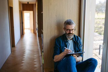 Man standing by the window with tablet and writing notes.