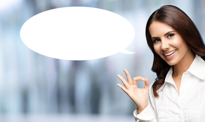 Portrait image - expressive happy smile business woman in white shirt show point ok okay hand sign gesture. Businesswoman stand on blurred office background with empty free space mockup speech bubble