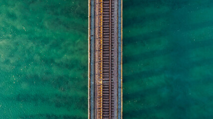 Top-down aerial view of a railroad track bridge crossing clear turquoise ocean water, featuring strong symmetry, minimal composition, and textured tropical sea surface © Bpato Media