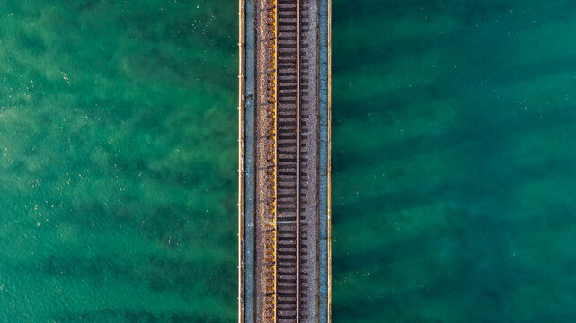 Top-down aerial view of a railroad track bridge crossing clear turquoise ocean water, featuring strong symmetry, minimal composition, and textured tropical sea surface