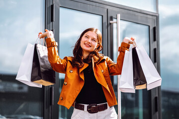 A woman with long hair smiles as she holds several shopping bags while standing outside a contemporary glass building on a sunny day. Consumer, fashion, shopping, shopping, lifestyle concept