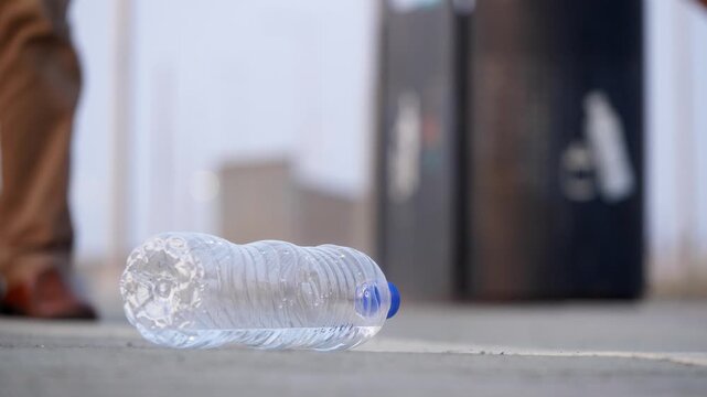 A close-up daylight outdoor shot of an empty plastic water bottle lying on a city road or sidewalk with a recyclable dust bin or garbage can, and a girl passing on a kids' kick scooter in background