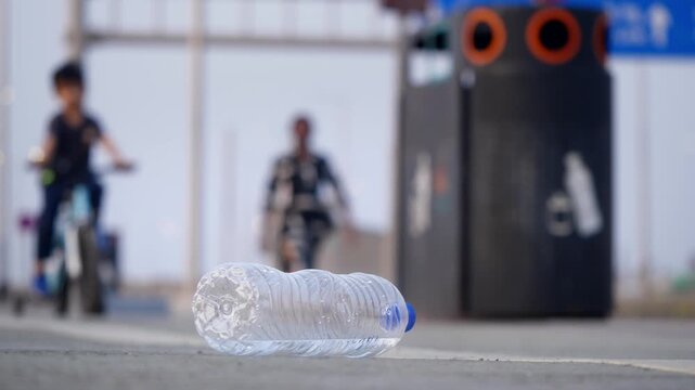A close-up daylight outdoor shot of an empty plastic water bottle lying on a city road or sidewalk with a recyclable dust bin or garbage can, and a male kid on a small bicycle passing in background.