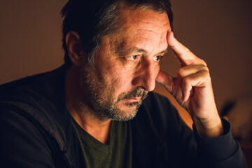 Concerned man looking at computer screen and thinking in dark room illuminated by warm low key...