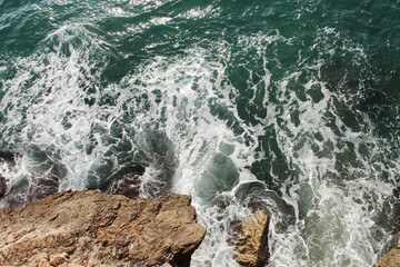 A high-angle aerial view of the powerful, turquoise, green, and white ocean waves with foam crashing forcefully against the dark, rugged, natural coastal rocks on a bright, sunny day