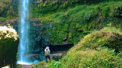 Adventurer in Hat and Backpack Admiring Hidden Waterfall and Mossy Cave in Lush Greenery at Ragia Forest in the Aberdare Ranges, Kenya