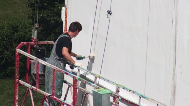 Male Worker In Overalls Applies White Paint To Facade With Roller.