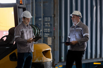 Two warehouse workers in uniform and helmets discuss logistics operations in front of large shipping container and forklift at loading dock.