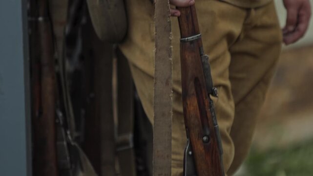 Red Army private soldier holds Mosin rifle, standing guard near display of weapons and army tents. Reenactment portrays field life of Great Patriotic War.