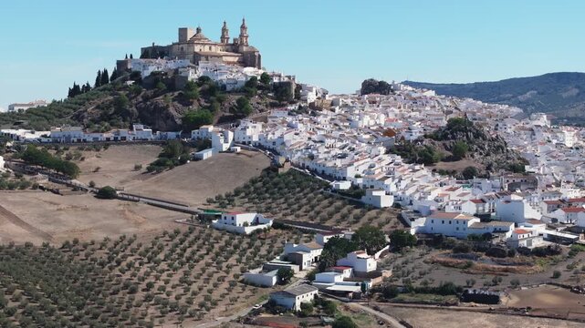 Wide aerial drone view of Olvera white hill town in Andalusia, Spain, featuring historic castle and church above a dense whitewashed village, with olive groves and countryside hills. Cinematic grading