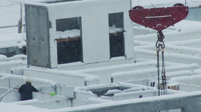 One person in dark workwear with yellow hard hat clearing fresh snow from concrete structures using green shovel. Active snowfall over industrial site creates harsh winter conditions.