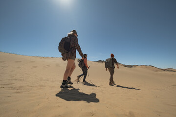 Hikers with backpacks and trekking poles walk single file along a sand dune in Mongolia. Side-view shot captures their steady pace from left to right under a clear summer sky.