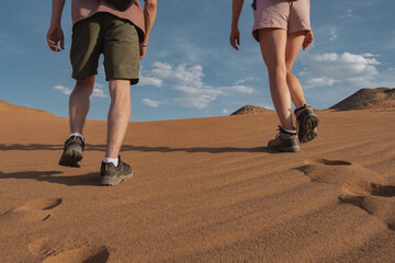 A couple in shorts with backpacks ascends a sand dune in the Mongolian desert. Low rear-angle medium shot captures their boot soles and footprints on the steep sandy slope.