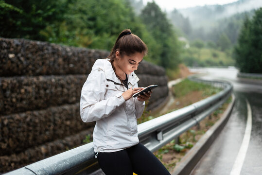 Girl in sportswear using smartphone on a rainy mountain road guardrail